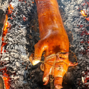 A whole pig, expertly roasted to a deep, glossy golden-brown. The skin appears exceptionally crispy and smooth, secured to a heavy metal rod with thin binding strings.