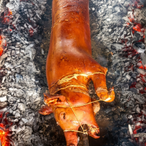 A whole pig, expertly roasted to a deep, glossy golden-brown. The skin appears exceptionally crispy and smooth, secured to a heavy metal rod with thin binding strings.