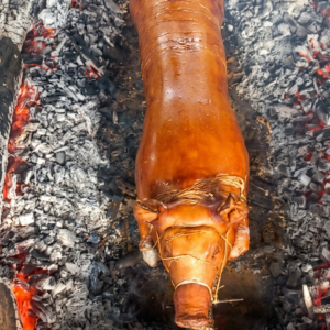 A whole pig, expertly roasted to a deep, glossy golden-brown. The skin appears exceptionally crispy and smooth, secured to a heavy metal rod with thin binding strings.