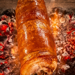 A whole, perfectly roasted lechon belly roll on a rotisserie spit, featuring a deeply caramelized, glistening golden-brown crackling skin, positioned over a bed of glowing red charcoal embers.